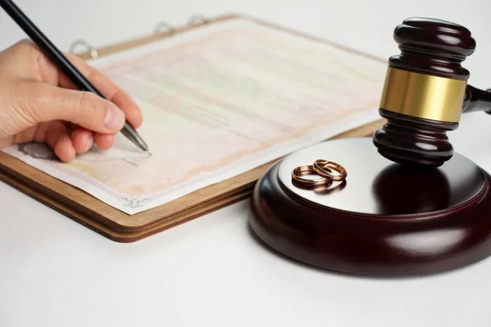 A person signing a legal document with a pair of wedding rings resting on a gavel, symbolizing divorce proceedings.