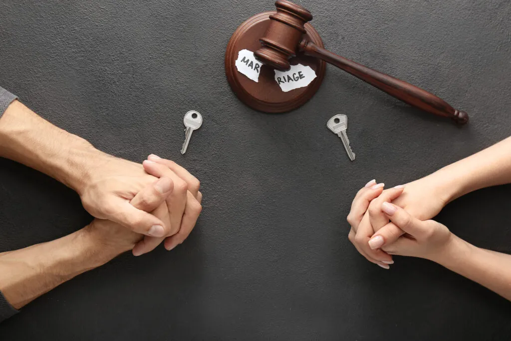 Two people sitting opposite each other at a table with keys and a gavel over a torn piece of paper, representing a divorce or legal separation.