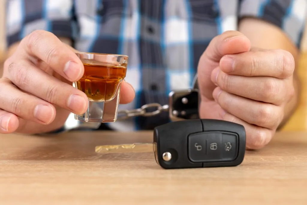 A pair of hands in handcuffs holding a shot of alcohol next to a car key, illustrating a DUI charge.