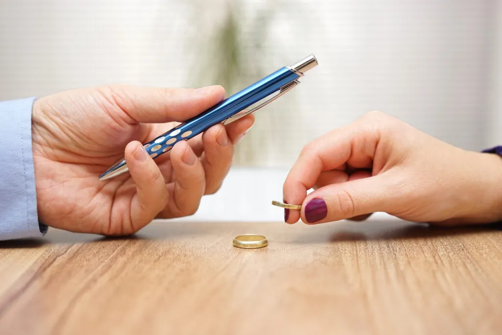 A hand holding a pen toward another hand holding a wedding ring over a table, representing a divorce settlement.