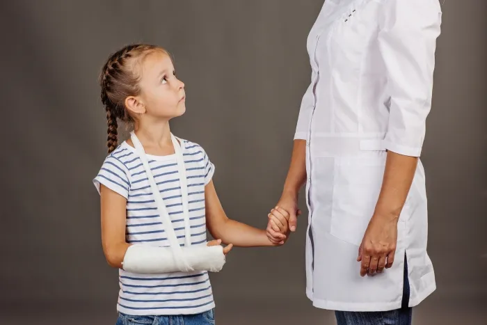 A young girl with a broken arm in a sling holding hands with a person wearing a medical coat.